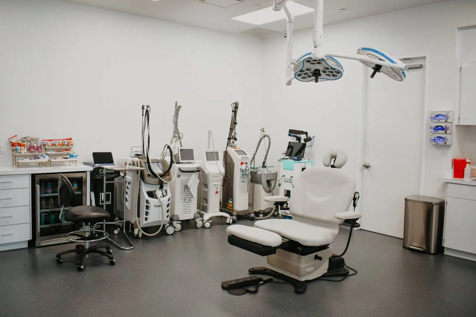 Dermatology treatment room with multiple laser and energy-based devices, examination chair, overhead surgical lights, and medical equipment at Regency Specialties